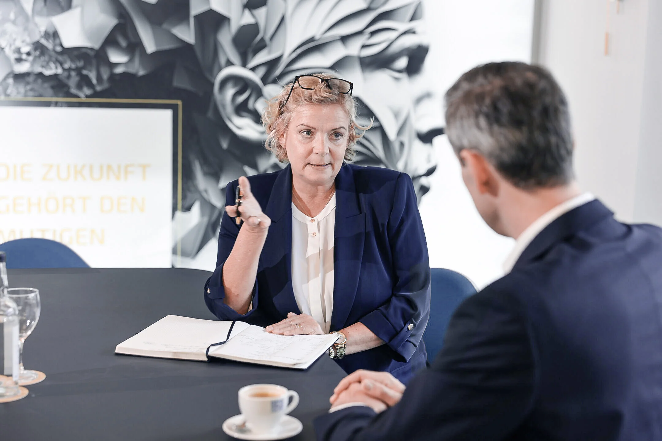 Beratungsgespräch bei Denkströme: Eine Führungskraft spricht am Konferenztisch und macht Notizen, gegenüber sitzt ein Gesprächspartner; Kaffeetasse und Wasser stehen bereit.