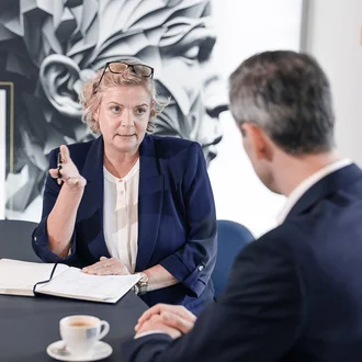 Beratungsgespräch bei Denkströme: Eine Führungskraft spricht am Konferenztisch und macht Notizen, gegenüber sitzt ein Gesprächspartner; Kaffeetasse und Wasser stehen bereit.