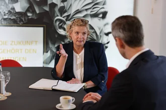 Beratungsgespräch bei Denkströme: Eine Führungskraft spricht am Konferenztisch und macht Notizen, gegenüber sitzt ein Gesprächspartner; Kaffeetasse und Wasser stehen bereit.