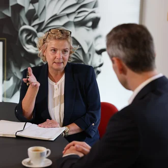 Beratungsgespräch bei Denkströme: Eine Führungskraft spricht am Konferenztisch und macht Notizen, gegenüber sitzt ein Gesprächspartner; Kaffeetasse und Wasser stehen bereit.