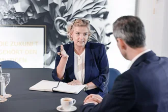 Beratungsgespräch bei Denkströme: Eine Führungskraft spricht am Konferenztisch und macht Notizen, gegenüber sitzt ein Gesprächspartner; Kaffeetasse und Wasser stehen bereit.