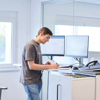 Junger Mann an einem modernen Arbeitsplatz mit zwei Monitoren beim Bearbeiten von Dokumenten – technisches Büro, Ausbildung im kaufmännischen Bereich, Arbeiten im Stehen