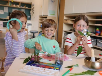 Drei Kinder malen und untersuchen Blätter mit Lupen in einem Gruppenraum der Kindertagesstätte