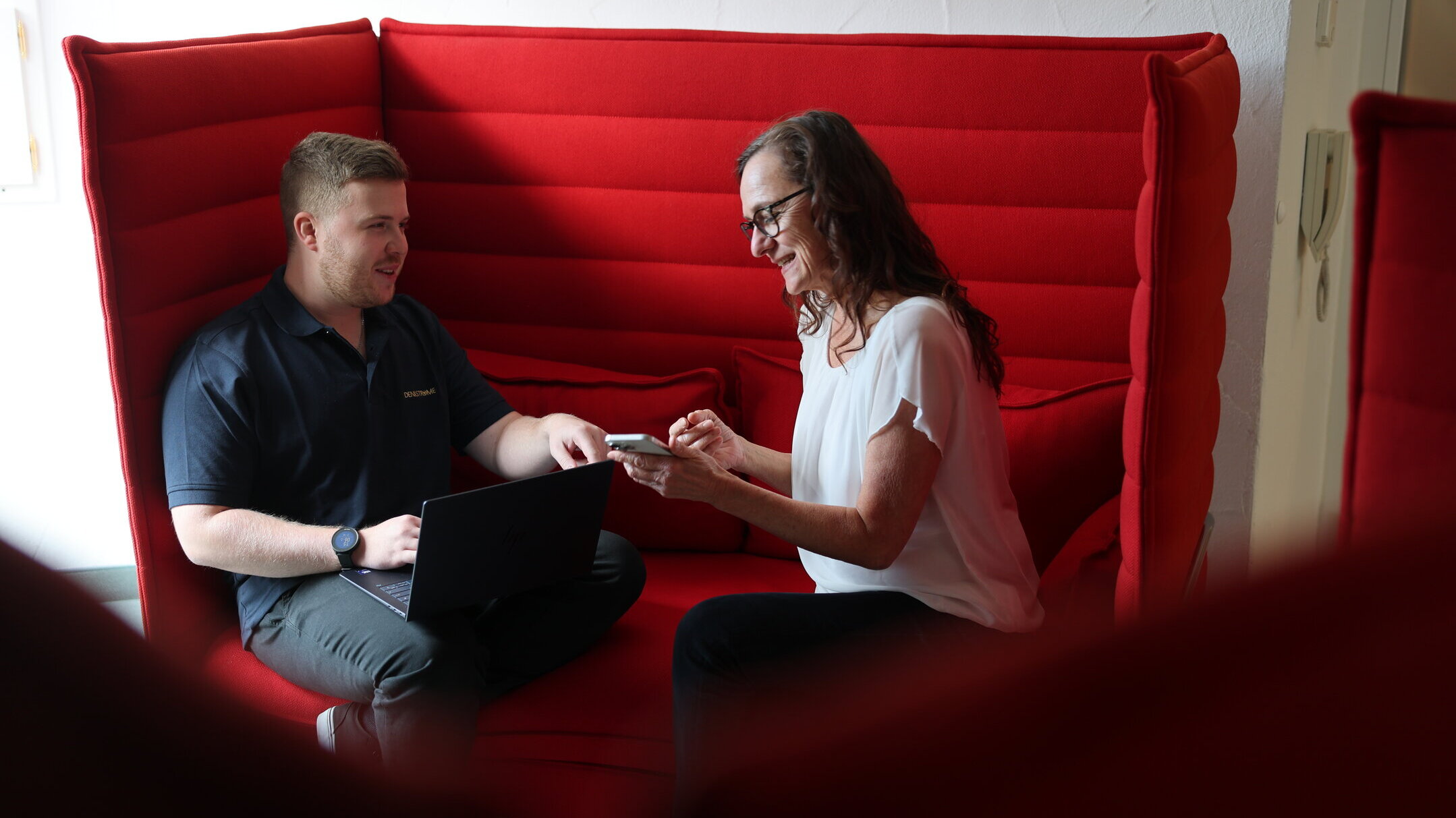 Zwei Mitarbeitende aus der Redaktion von Denkströme sitzen in einer roten Sitznische und besprechen Inhalte; Laptop und Smartphone liegen bereit.