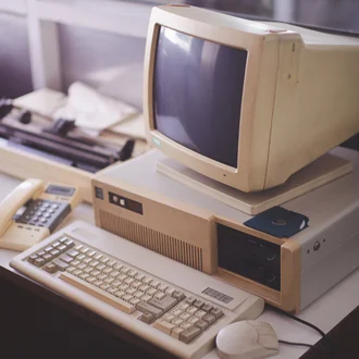 Alter Desktop-Computer mit Röhrenmonitor, Tastatur, Maus, Floppy-Disk, Nadeldrucker und Festnetztelefon auf einem Schreibtisch – Retro-Technologie aus den 1980er-Jahren