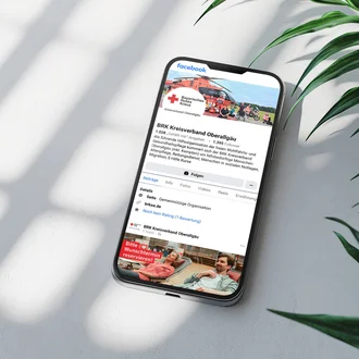 Smartphone auf weißem Tisch mit geöffneter Facebook-Seite des BRK Kreisverbands Oberallgäu – digitale Öffentlichkeitsarbeit und Social-Media-Präsenz des Bayerischen Roten Kreuzes