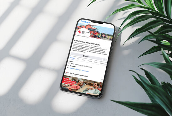 Smartphone auf weißem Tisch mit geöffneter Facebook-Seite des BRK Kreisverbands Oberallgäu – digitale Öffentlichkeitsarbeit und Social-Media-Präsenz des Bayerischen Roten Kreuzes