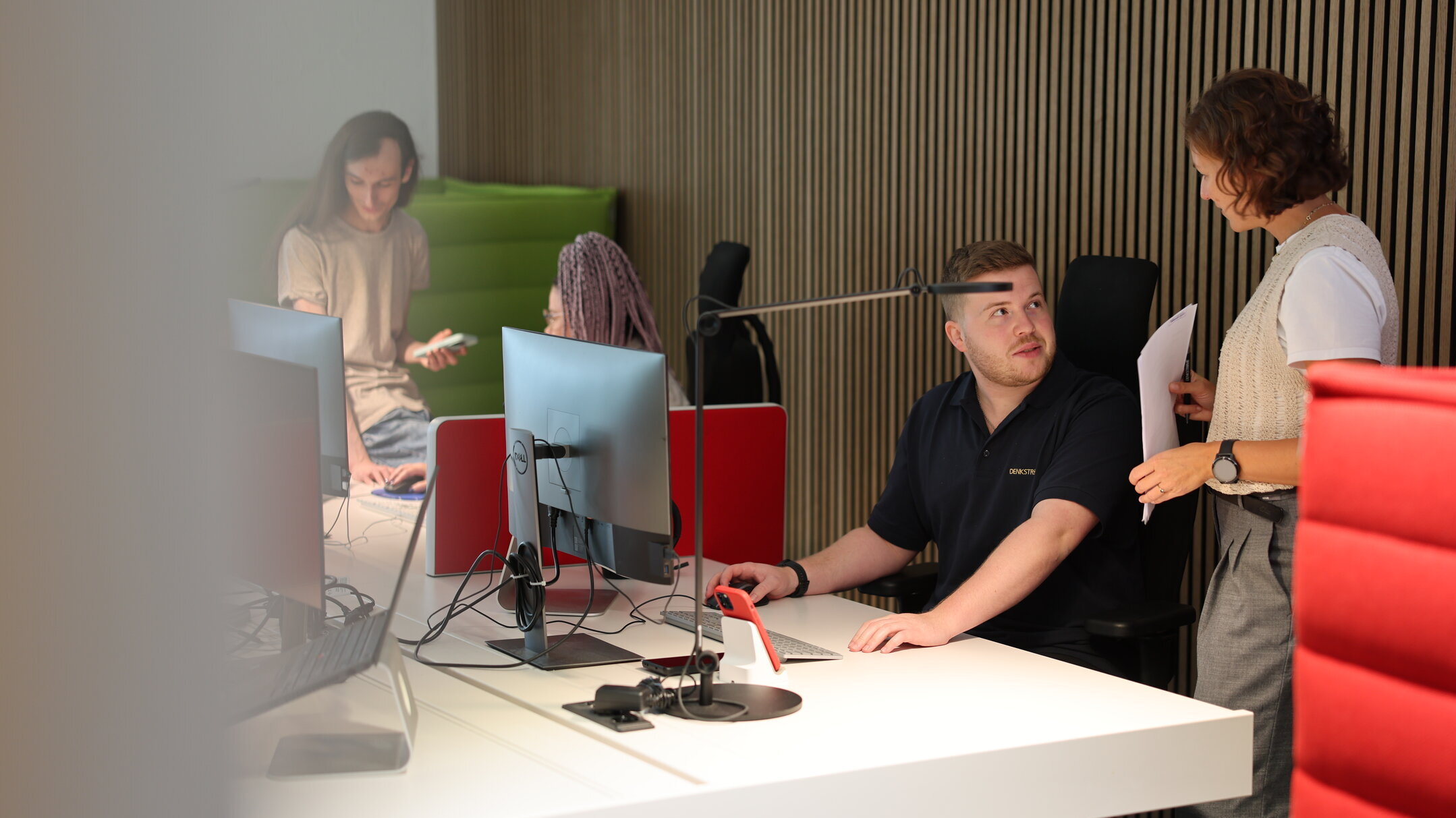 Marketing-Outsourcing bei Denkströme: Mitarbeitende arbeiten an Desktop-PCs im Büro; eine Kollegin bespricht Unterlagen mit einem sitzenden Teammitglied am Arbeitsplatz.