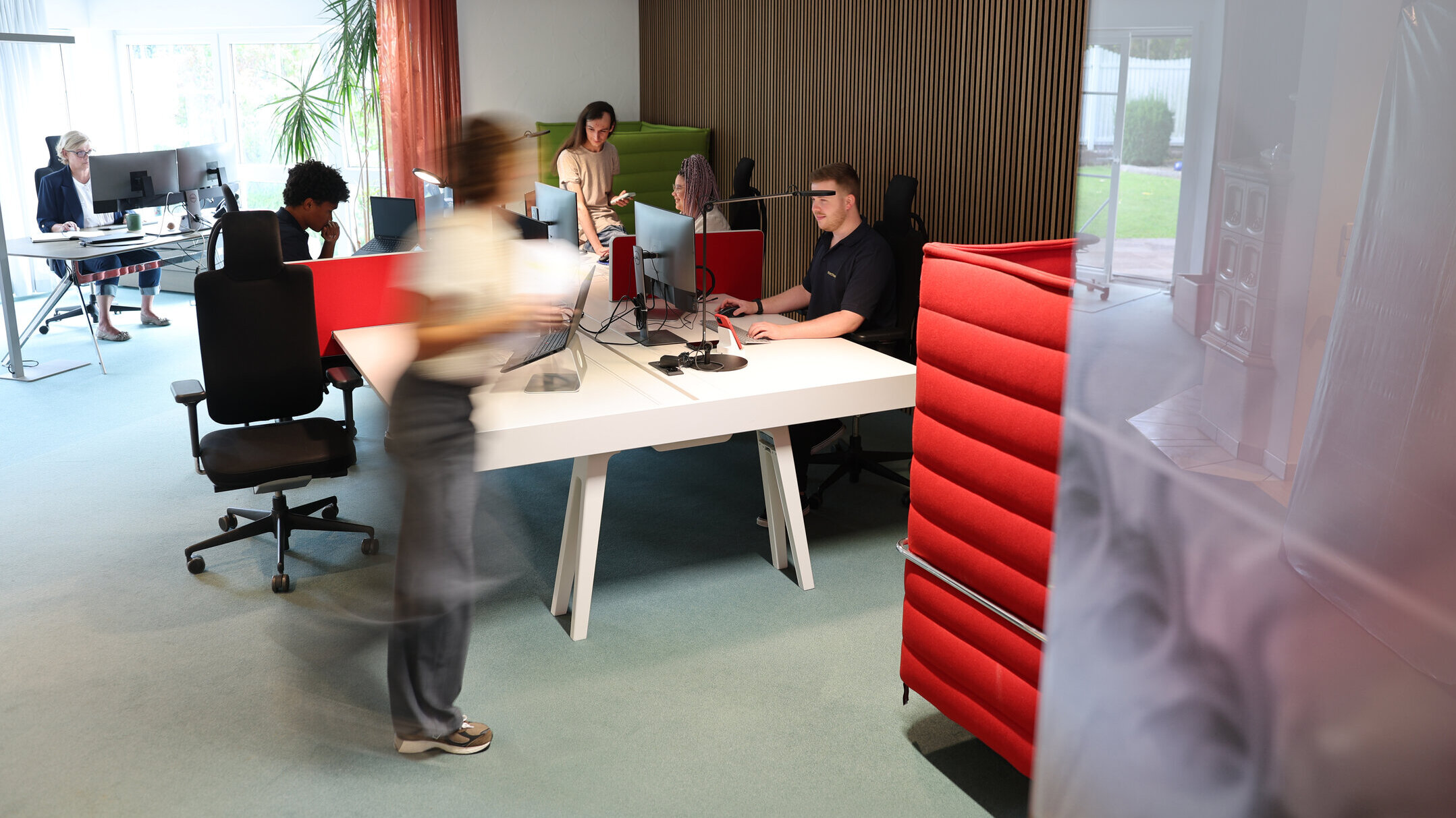 Offenes Büro bei Denkströme in Kempten: Mitarbeitende arbeiten an Schreibtischen mit Monitoren, eine Person bewegt sich durch den Raum; moderne Arbeitsplätze mit roten Akustikelementen.