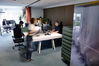 Offenes Büro bei Denkströme in Kempten: Mitarbeitende arbeiten an Schreibtischen mit Monitoren, eine Person bewegt sich durch den Raum; moderne Arbeitsplätze mit roten Akustikelementen.