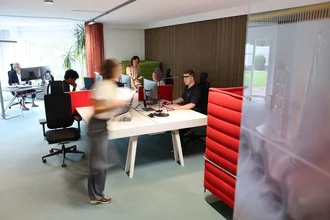 Offenes Büro bei Denkströme in Kempten: Mitarbeitende arbeiten an Schreibtischen mit Monitoren, eine Person bewegt sich durch den Raum; moderne Arbeitsplätze mit roten Akustikelementen.