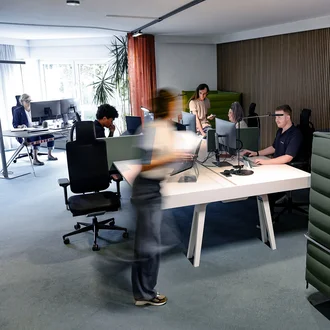 Offenes Büro bei Denkströme in Kempten: Mitarbeitende arbeiten an Schreibtischen mit Monitoren, eine Person bewegt sich durch den Raum; moderne Arbeitsplätze mit roten Akustikelementen.