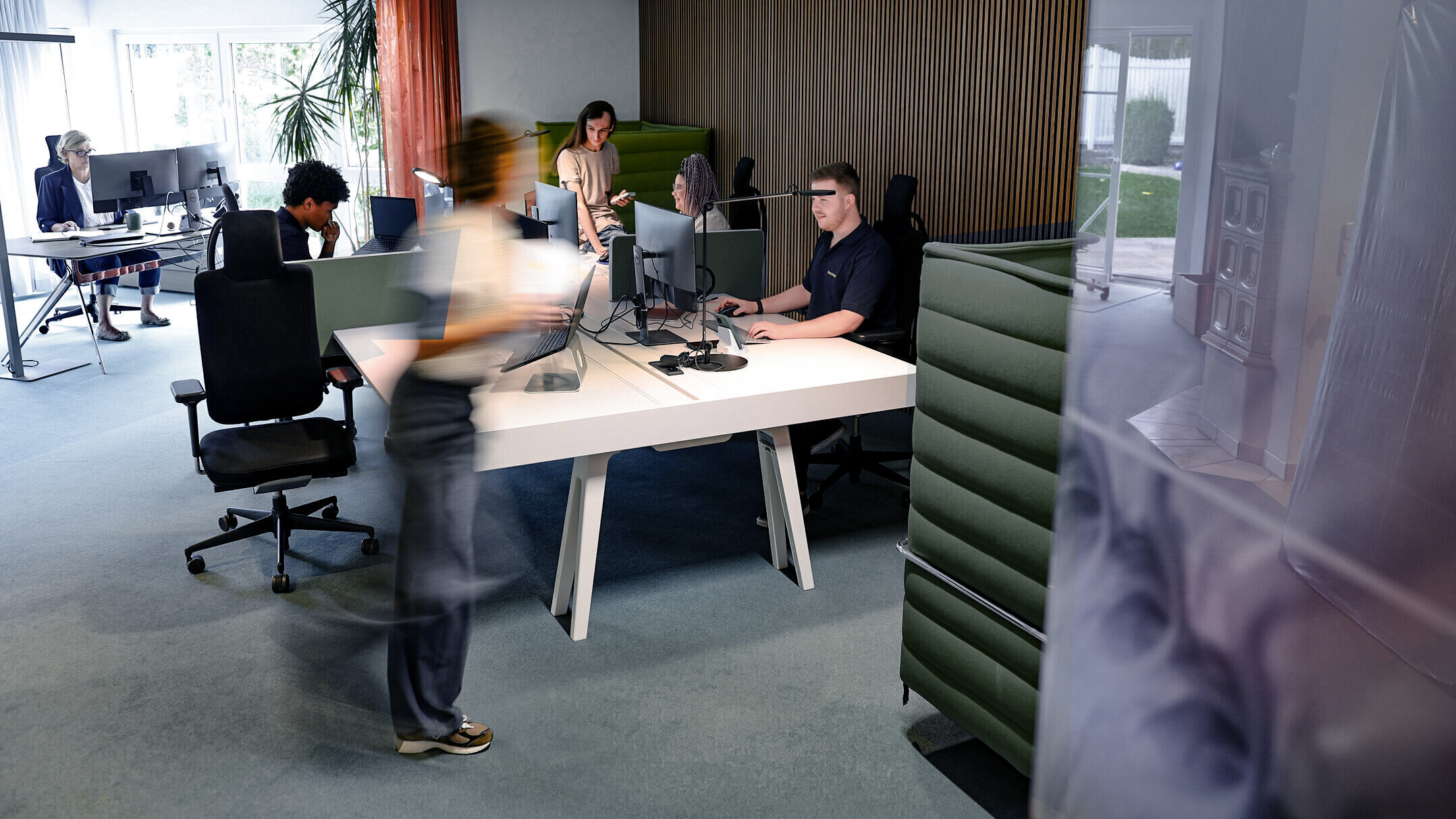 Offenes Büro bei Denkströme in Kempten: Mitarbeitende arbeiten an Schreibtischen mit Monitoren, eine Person bewegt sich durch den Raum; moderne Arbeitsplätze mit roten Akustikelementen.