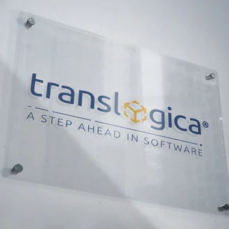 Transparentes Acrylschild mit dem translogica-Logo und Slogan „A step ahead in software“, montiert an einer hellen Wand – moderner, professioneller Markenauftritt eines Softwareunternehmens mit Fokus auf Innovation und Logistiklösungen.