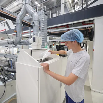 Junger Mitarbeiter mit Haarnetz füllt weißen Kunststoffgranulat in eine industrielle Verarbeitungsmaschine – moderne Produktionshalle mit Rohren, Technik und Kontrollsystemen im Hintergrund