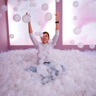 Frau spielt lachend in einem rosa Bällebad voller weißer Kugeln vor kreativem Hintergrund 