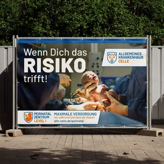 Bauzaunbanner des Allgemeinen Krankenhauses Celle mit der Botschaft „Wenn Dich das Risiko trifft!“ – Werbung für das Perinatalzentrum Level 1 mit Bild einer Geburt und Informationen zur maximalen Versorgung bei Risikoschwangerschaften