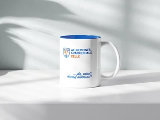 Weiße Kaffeetasse mit blauem Innenrand, Logo des Allgemeinen Krankenhauses Celle und Slogan „... da, wenn’s darauf ankommt!“ auf heller Fläche