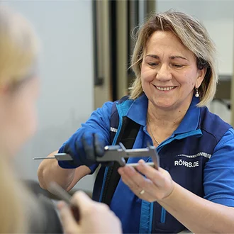 Mitarbeiterin der Dr. Werner Röhrs GmbH & Co. KG in blauer Arbeitskleidung zeigt einer jungen Frau die Anwendung eines Messschiebers im technischen Umfeld – Szene in einem modernen Labor oder Qualitätsprüfraum mit Fokus auf Präzision und Ausbildung.