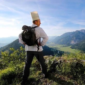 Koch mit Rucksack und Berufsbekleidung schaut ins Alpenpanorama – Symbolbild für Beruf und Berufung in Einklang, Teil der Arbeitgeberkampagne der Fachklinik Allgäu.