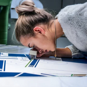 Mitarbeiterin prüft sorgfältig einen Druckbogen mit Lupe – Detailaufnahme eines kreativen Produktionsprozesses im Bereich Markenkreation bei der Denkströme