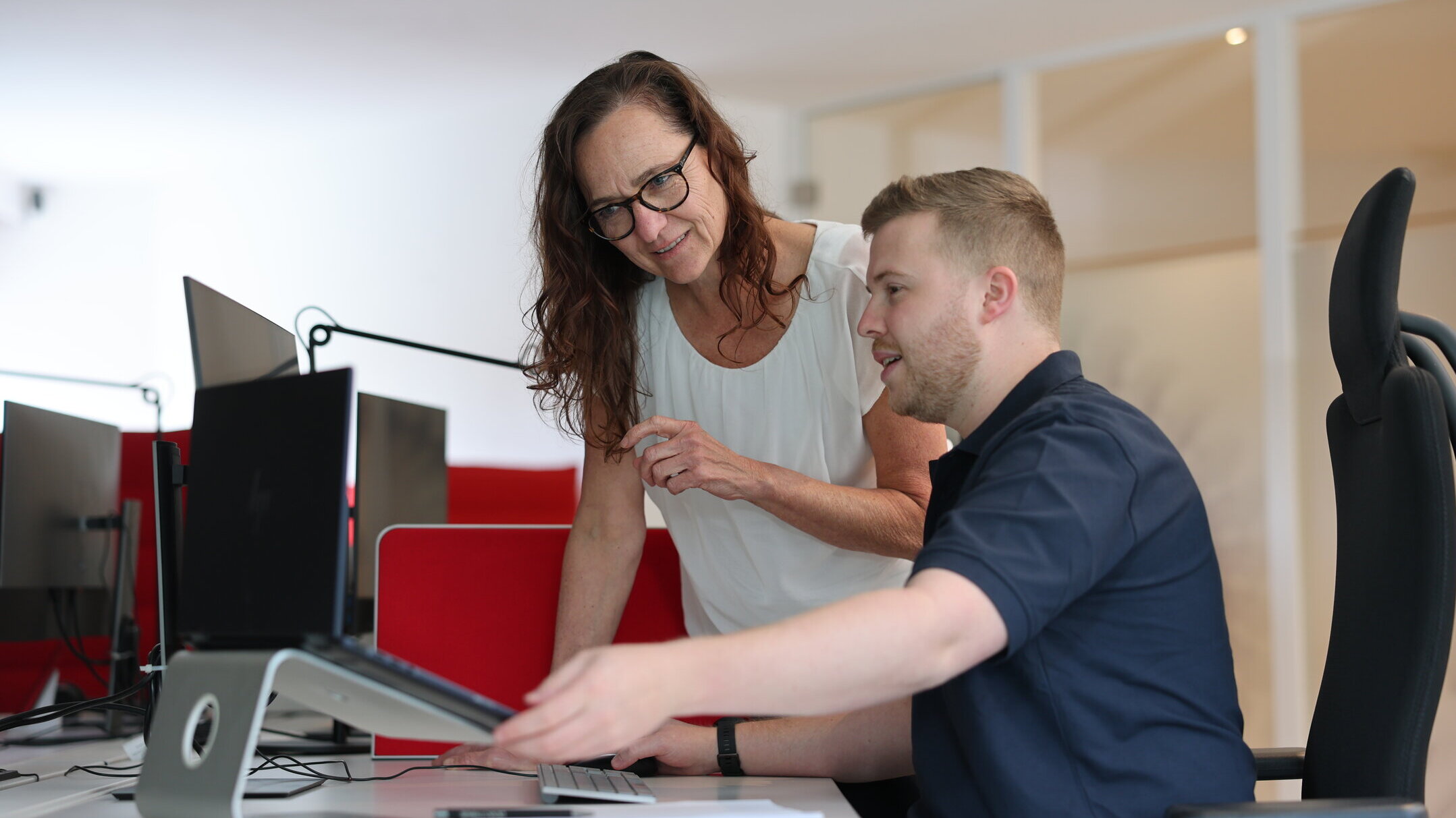 Zwei Mitarbeitende aus der Redaktion von Denkströme arbeiten am Schreibtisch: Eine Person zeigt auf den Monitor, die andere sitzt am Computer und hört zu.