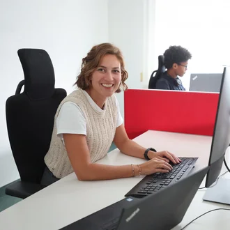 Mitarbeiterin bei Denkströme arbeitet am Schreibtisch am Computer und lächelt in die Kamera; im Hintergrund sitzt ein Kollege an einem weiteren Arbeitsplatz.