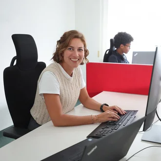 Mitarbeiterin bei Denkströme arbeitet am Schreibtisch am Computer und lächelt in die Kamera; im Hintergrund sitzt ein Kollege an einem weiteren Arbeitsplatz.