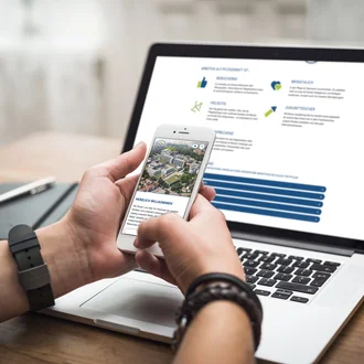 Person bedient Smartphone mit Startseite des Klinikverbunds Allgäu vor geöffnetem Laptop mit Karriereseite zur Pflegeausbildung.