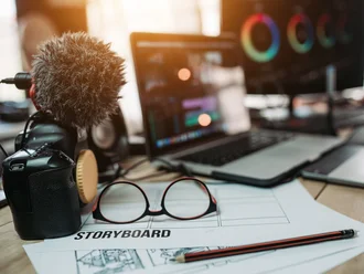 Kamera mit Mikrofon, Storyboard, Brille und Laptop mit geöffneter Videoschnittsoftware auf einem Schreibtisch – kreative Arbeitsumgebung für Videoproduktion