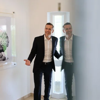 Eingangsbereich bei Denkströme: Ein Mann im Anzug steht lächelnd in der Tür, daneben hängt ein Bild mit stilisiertem Baum; seine Spiegelung ist in einer Glasfläche zu sehen.