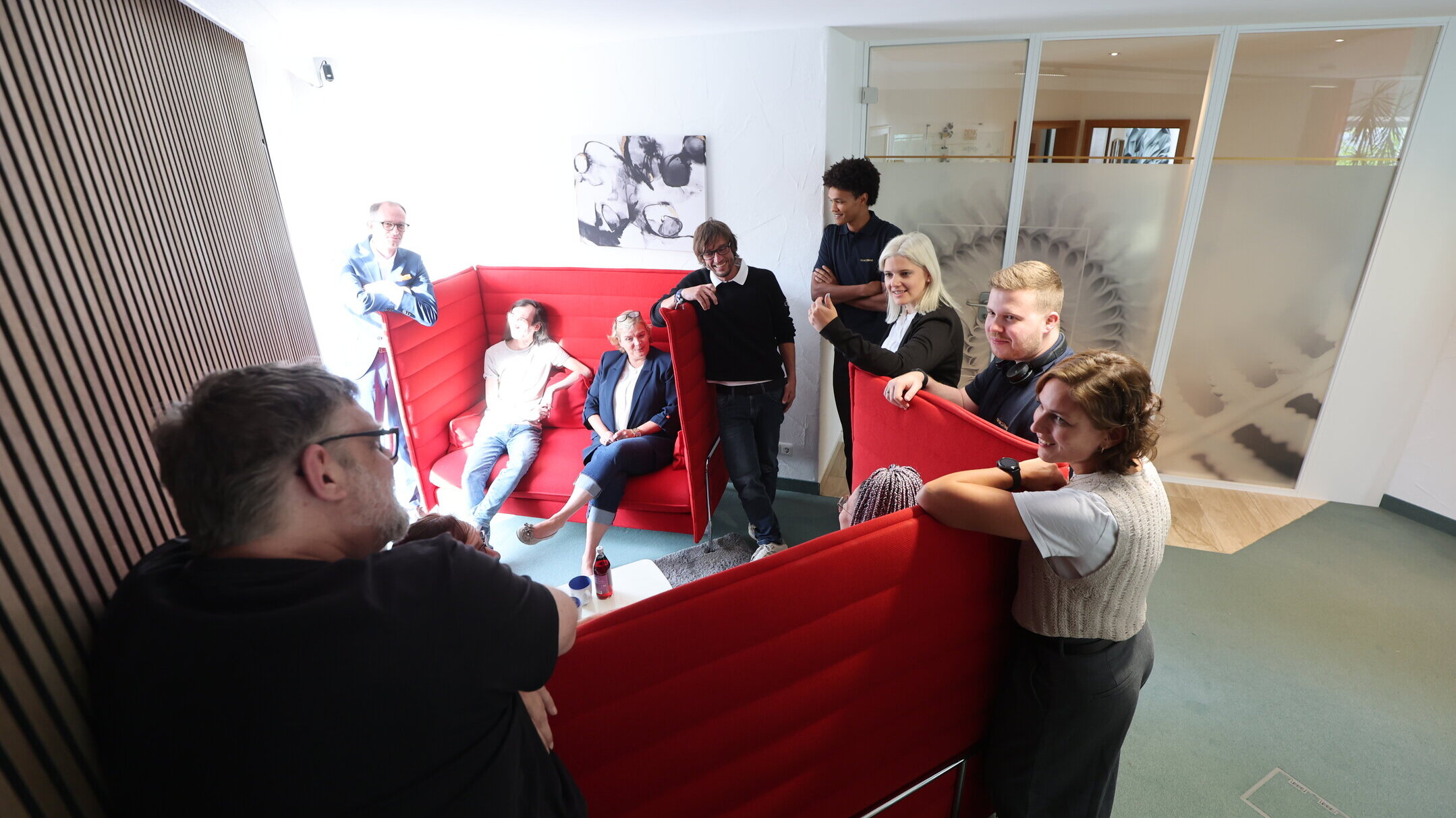 Team der Denkströme im Büro: Mitarbeitende stehen und sitzen in roten Akustik-Sitznischen und tauschen sich in einem hellen Aufenthaltsbereich aus