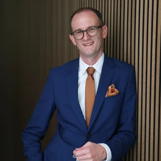 Fabian Schmid, leitender Geschäftsführer von Denkströme, steht im blauen Anzug vor einer Holzlamellenwand und lächelt in die Kamera.