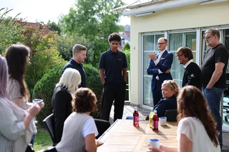 Team von Denkströme auf der Terrasse am Standort Kempten: Mitarbeitende stehen und sitzen um einen Tisch, sprechen miteinander und trinken Kaffee im Gartenbereich.