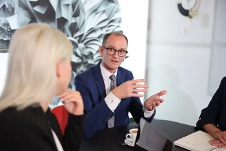 Geschäftsleitung von Denkströme im Meeting: Führungskraft im blauen Anzug spricht und gestikuliert am Besprechungstisch, Kolleginnen hören zu; Laptop und Notizen liegen bereit.