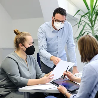 Teammeeting im Büro des Deutschen Herzzentrums München– drei Mitarbeitende mit FFP2-Masken im Gespräch über Unterlagen und Tablet am Arbeitsplatz