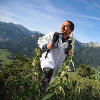 Lächelnder Arzt in weißem Kittel und mit Rucksack auf Wanderung durch die Alpen – Symbolbild für medizinische Karriere mit Lebensqualität bei der Fachklinik Allgäu.