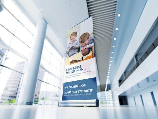 Roll-up-Banner des Klinikverbund Allgäu zur Azubi-Kampagne mit dem Slogan „Jeder fängt mal klein an“, abgebildete Kinder in Pflege-Rollenspielen, Call-to-Action mit Bewerbungslink für Ausbildungsplätze in der Gesundheitsbranche.