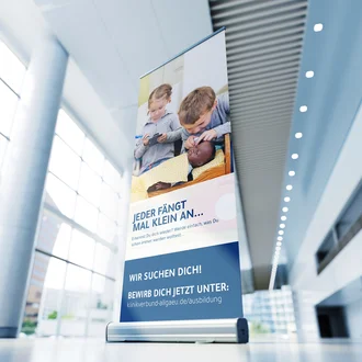 Roll-up-Banner des Klinikverbund Allgäu zur Azubi-Kampagne mit dem Slogan „Jeder fängt mal klein an“, abgebildete Kinder in Pflege-Rollenspielen, Call-to-Action mit Bewerbungslink für Ausbildungsplätze in der Gesundheitsbranche.