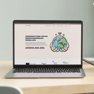Laptop auf einem modernen Holztisch zeigt die Startseite des Projekts „Regi-OA – Gemeinsam stark für ein widerstandsfähiges Oberallgäu“ mit gezeichneter Weltkugel in schützenden Händen als Symbol für Resilienz und Krisenvorsorge.