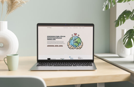 Laptop auf einem modernen Holztisch zeigt die Startseite des Projekts „Regi-OA – Gemeinsam stark für ein widerstandsfähiges Oberallgäu“ mit gezeichneter Weltkugel in schützenden Händen als Symbol für Resilienz und Krisenvorsorge.