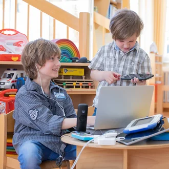Zwei Kinder sitzen am Spieltisch mit Laptop, Headset und Technikspielzeug – witziger Blick in den Berufsalltag von IT-Fachkräften im Klinikverbund Allgäu.