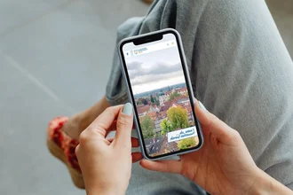 Frau hält Smartphone mit geöffneter Website der Stadt Celle in der Hand – TYPO3-optimierte Darstellung kommunaler Inhalte auf Mobilgerät mit Blick über die Dächer der Stadt