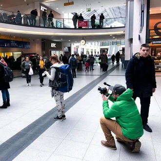 Kameramann im grünen Parka filmt junge Frau mit Rucksack in belebtem Bahnhofsgebäude mit Passanten und Geschäften im Hintergrund.