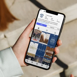Person hält Smartphone in der Hand und betrachtet das Instagram-Profil von „Dr. Werner Röhrs GmbH & Co. KG“, einem Hersteller für technische Federn, mit mehreren Beiträgen über Produkte, Mitarbeiter und Firmeninformationen – aufgenommen bei Tageslicht auf einer Steintreppe.