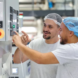 Zwei junge Männer mit Haarnetz und Arbeitskleidung bedienen gemeinsam ein modernes Steuerungspult in einer Produktionshalle – Teamarbeit und technische Ausbildung in der Industrie