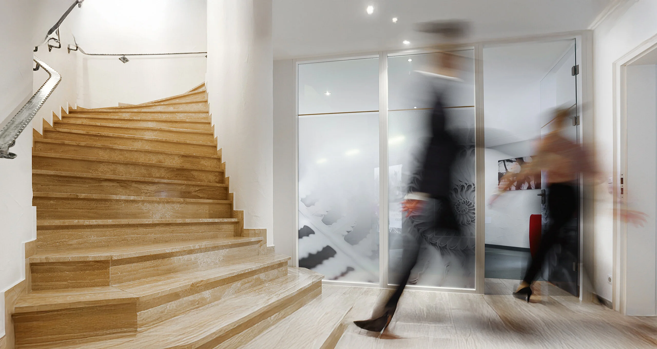 Flur im Denkströme-Büro mit breiter Treppe und Glaswand; zwei Personen gehen unscharf durch den Eingangsbereich.