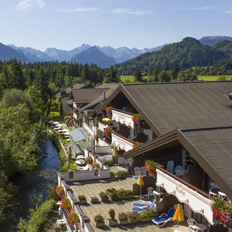 Hotelterrasse mit Liegestühlen am Flusslauf in den bayerischen Alpen bei Sonnenschein und Panoramablick auf bewaldete Berge