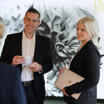 Projektmanagement bei Denkströme: Zwei Mitarbeitende im Gespräch im Büro, eine Person hält eine Kaffeetasse, die andere ein Laptop; Denkströme-Logo im Hintergrund.