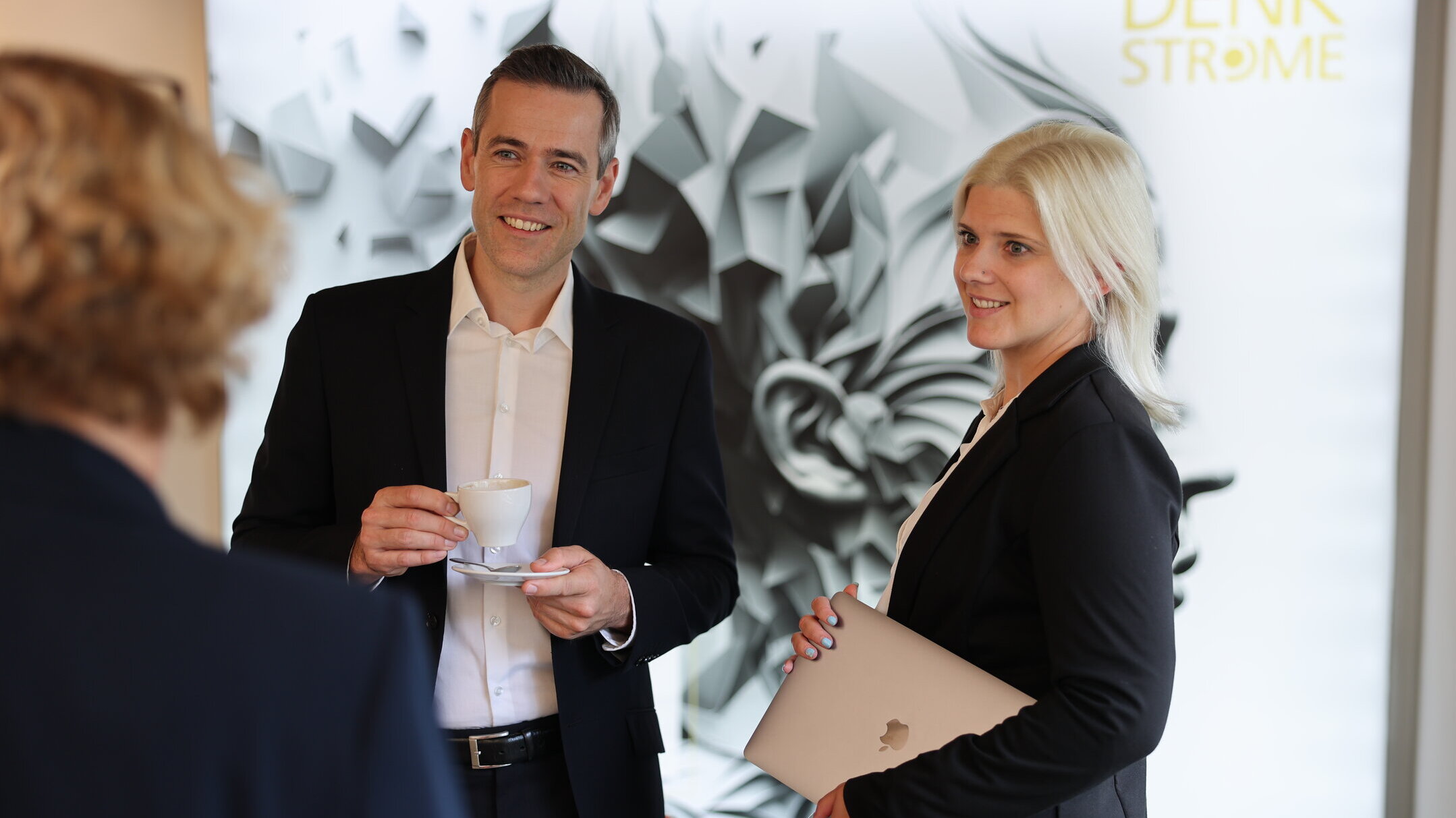 Projektmanagement bei Denkströme: Zwei Mitarbeitende im Gespräch im Büro, eine Person hält eine Kaffeetasse, die andere ein Laptop; Denkströme-Logo im Hintergrund.